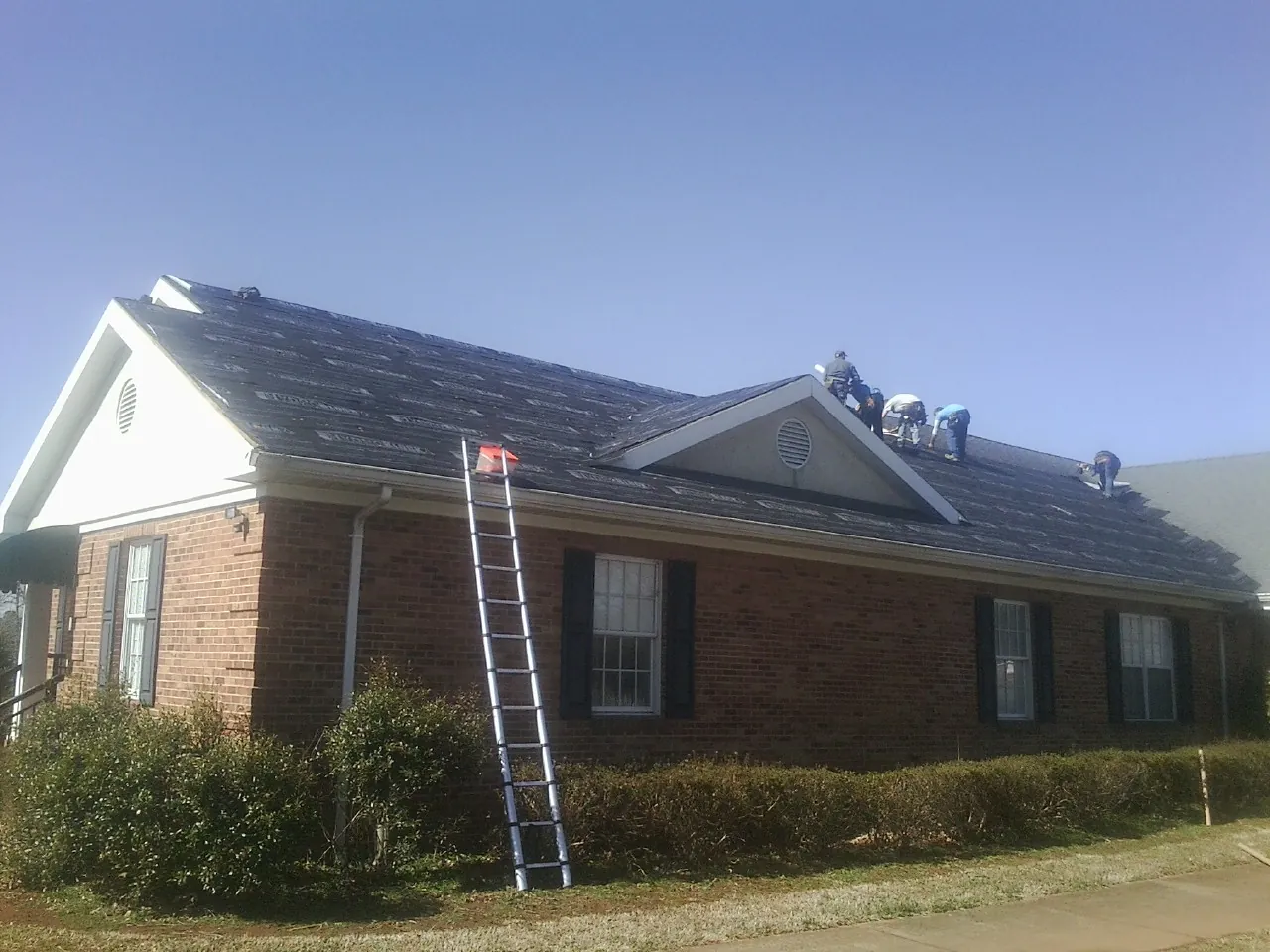 Crew tearing shingles off a brick ranch roof with a ladder set — Metal Roof Repair work by our crew in Brighton